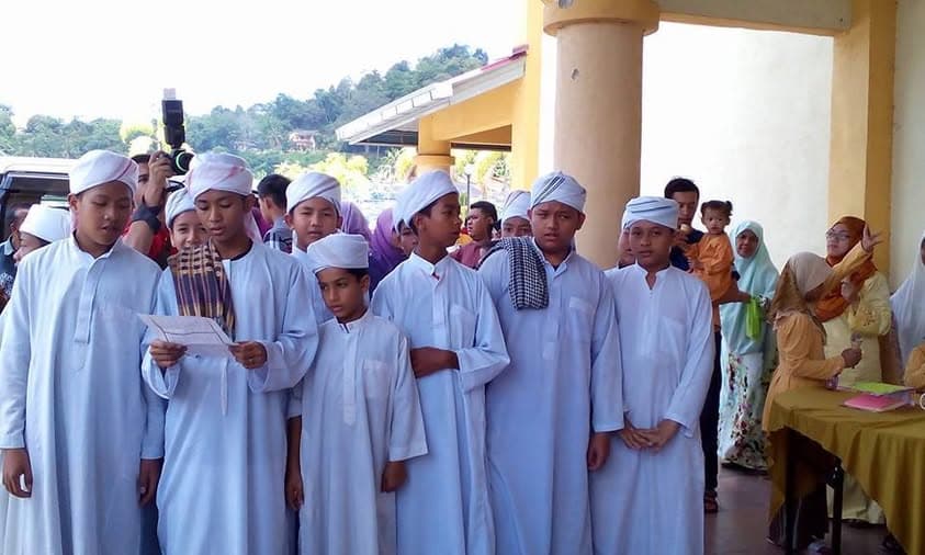 Sekumpulan pelajar berdiri bersama di sebuah majlis, seorang daripada mereka membaca daripada sekeping kertas